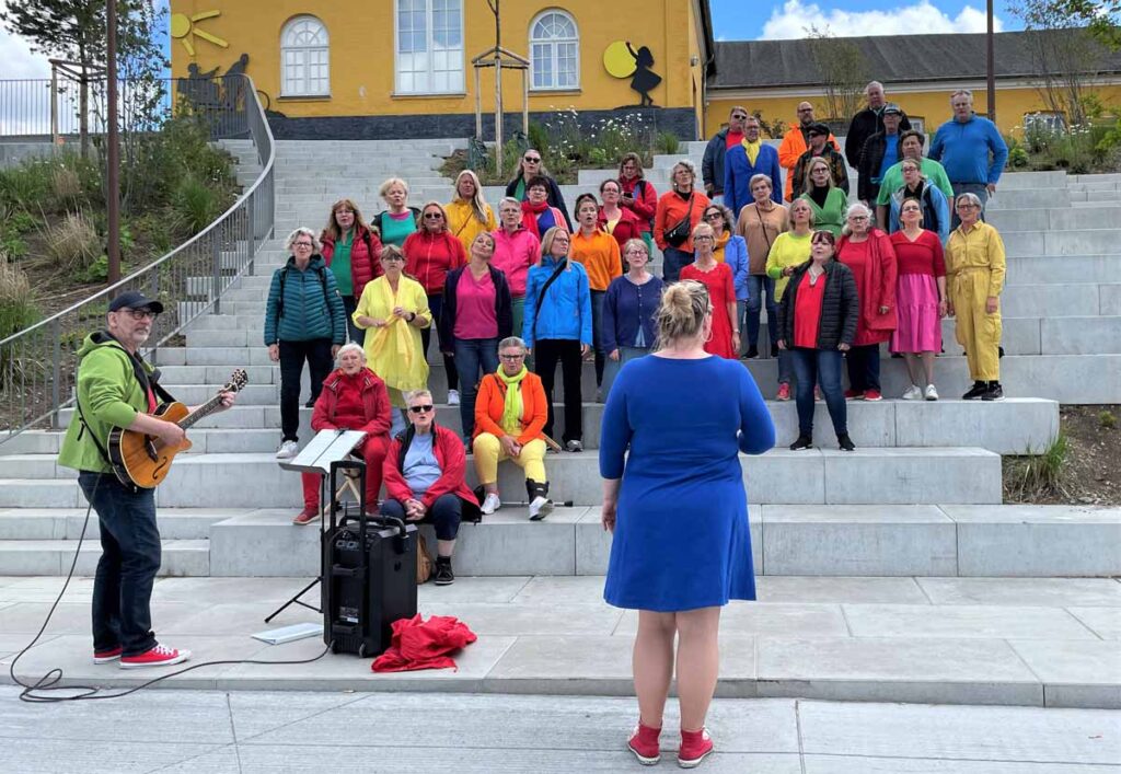 En del af koret ses på trappen på Kvægtorvet, Næstved klædt i spraglede farver med korleder Lonnie og Jens på guitar i forgrunden.